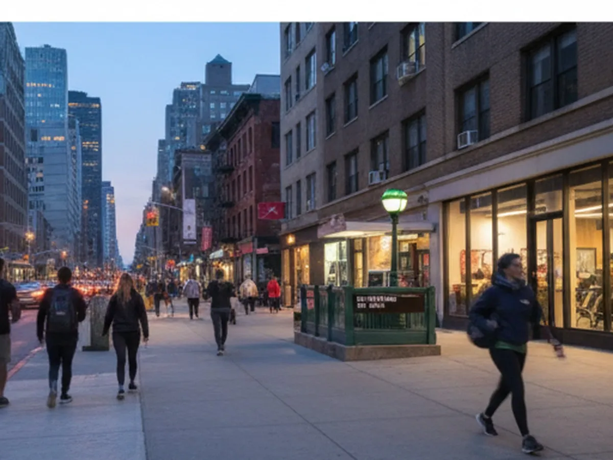 Twilight street-level view of a busy New York City avenue with a wide sidewalk, subway entrance, and pedestrians in athleisure walking past a brightly lit gym storefront, yellow taxis blurred in motion and mid-rise buildings framing the scene.