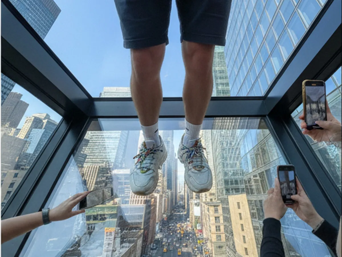 Daytime interior photo of a visitor stepping onto the glass skybox overlooking Madison Avenue, their sneakers visible against the clear floor, with taxis and pedestrians far below, nearby guests taking photos, and One Vanderbilt’s architectural details framing a bright blue sky.