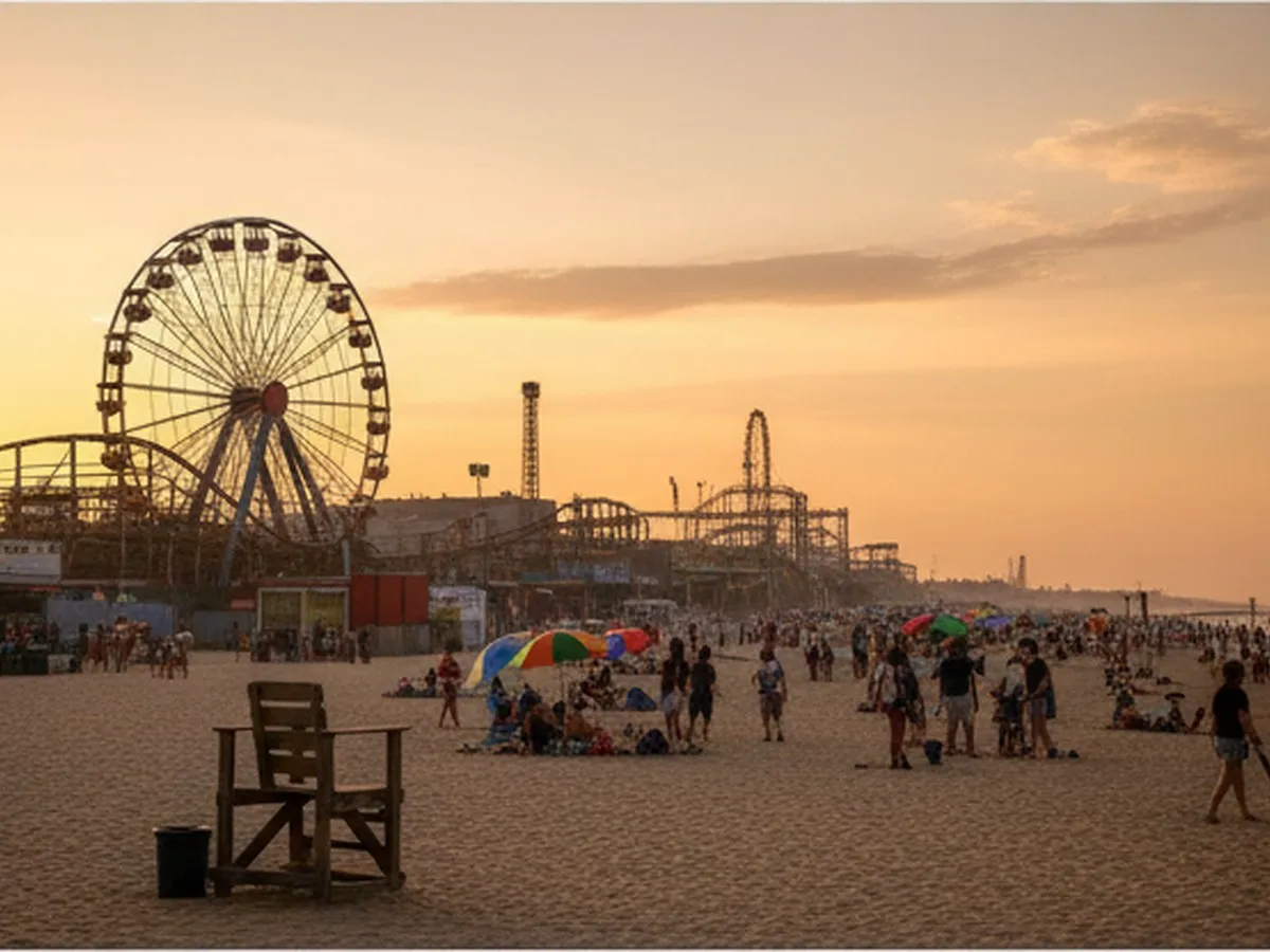 Late afternoon scene on Coney Island beach looking back toward the amusement district, Cyclone coaster and Wonder Wheel silhouetted against sky, scattered beachgoers and umbrellas, lifeguard chair in midground, soft warm light hinting the end of a busy holiday day.