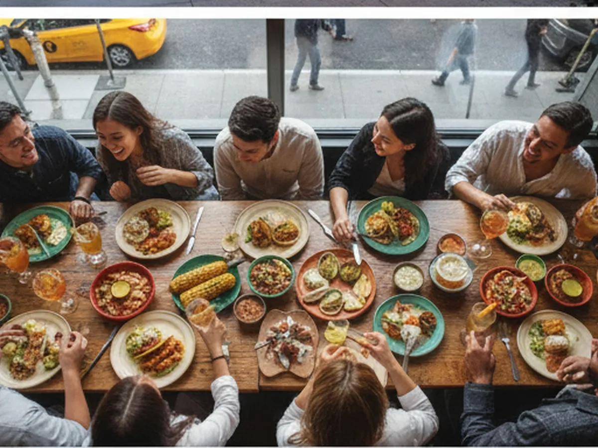 Overhead shot of a long wooden table in a lively NYC restaurant filled with shared Mexican dishes—tacos, mole, grilled corn, and mezcal cocktails—surrounded by a mixed group of young adults laughing, with blurred city lights visible through large windows.