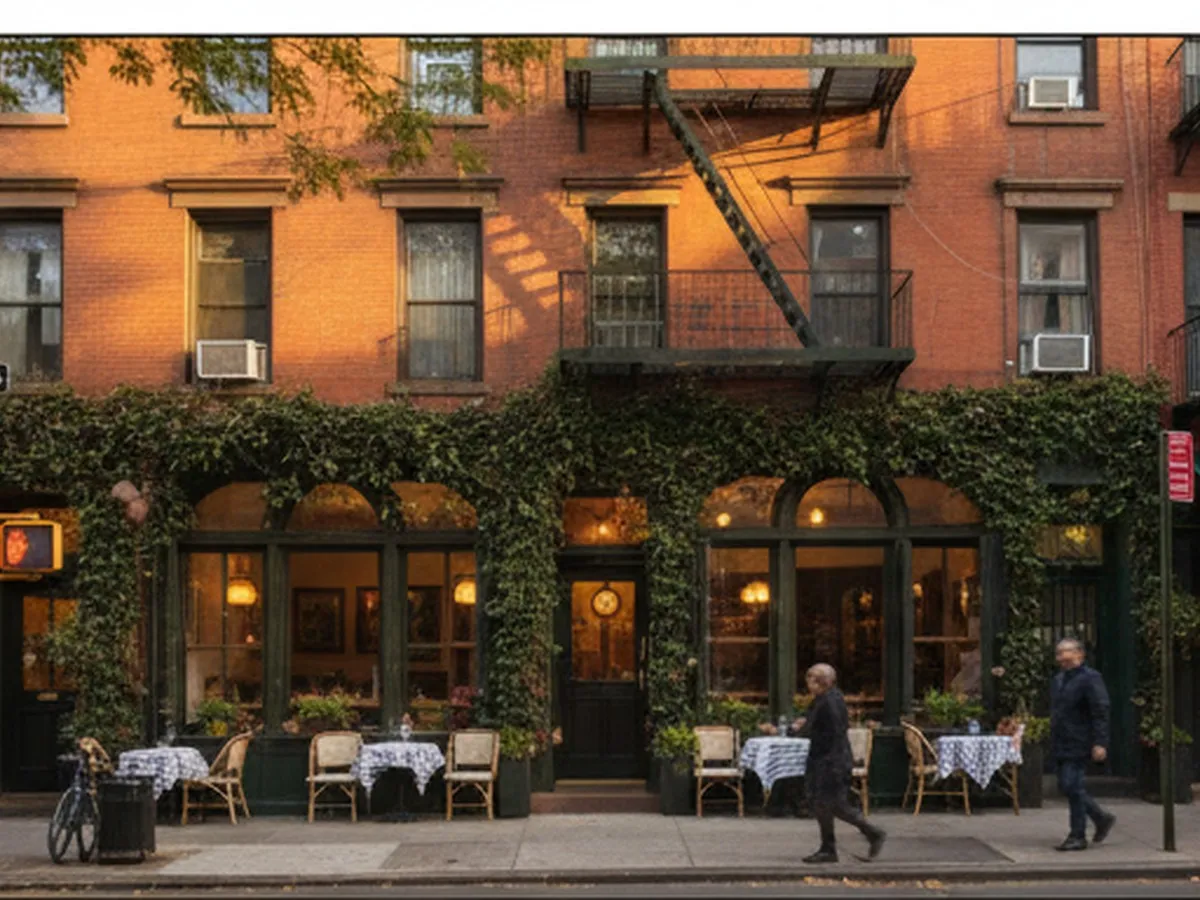 Golden-hour street view of Via Carota’s ivy-framed facade in Greenwich Village, with warm light spilling from tall windows, small bistro tables on the sidewalk, and New Yorkers walking past brownstone-style buildings and tree-lined West Village streets.