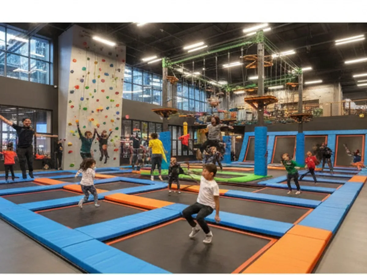 Interior scene of an NYC indoor trampoline and adventure park with adults and kids mid-jump on trampolines, a climbing wall in the background, and soft, even lighting that feels energetic but realistic.