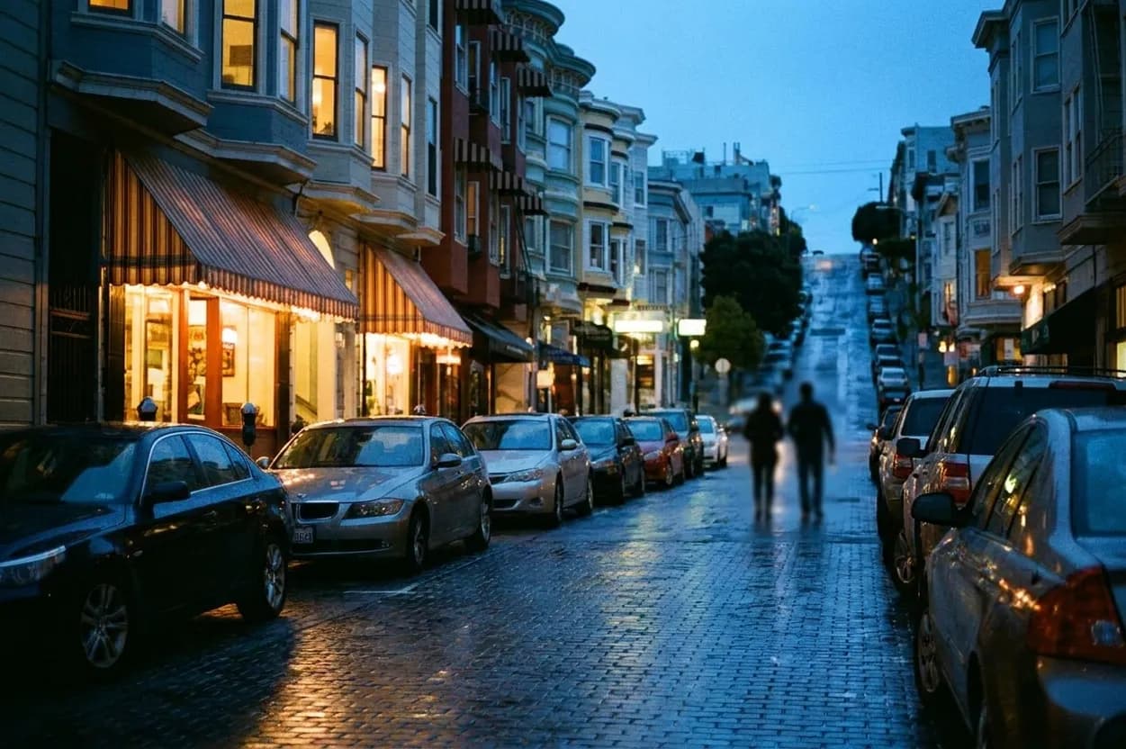 A narrow North Beach street in San Francisco at blue hour, with lit awnings over small storefronts, parked cars along a wet cobbled road, and a steep hill in the distance