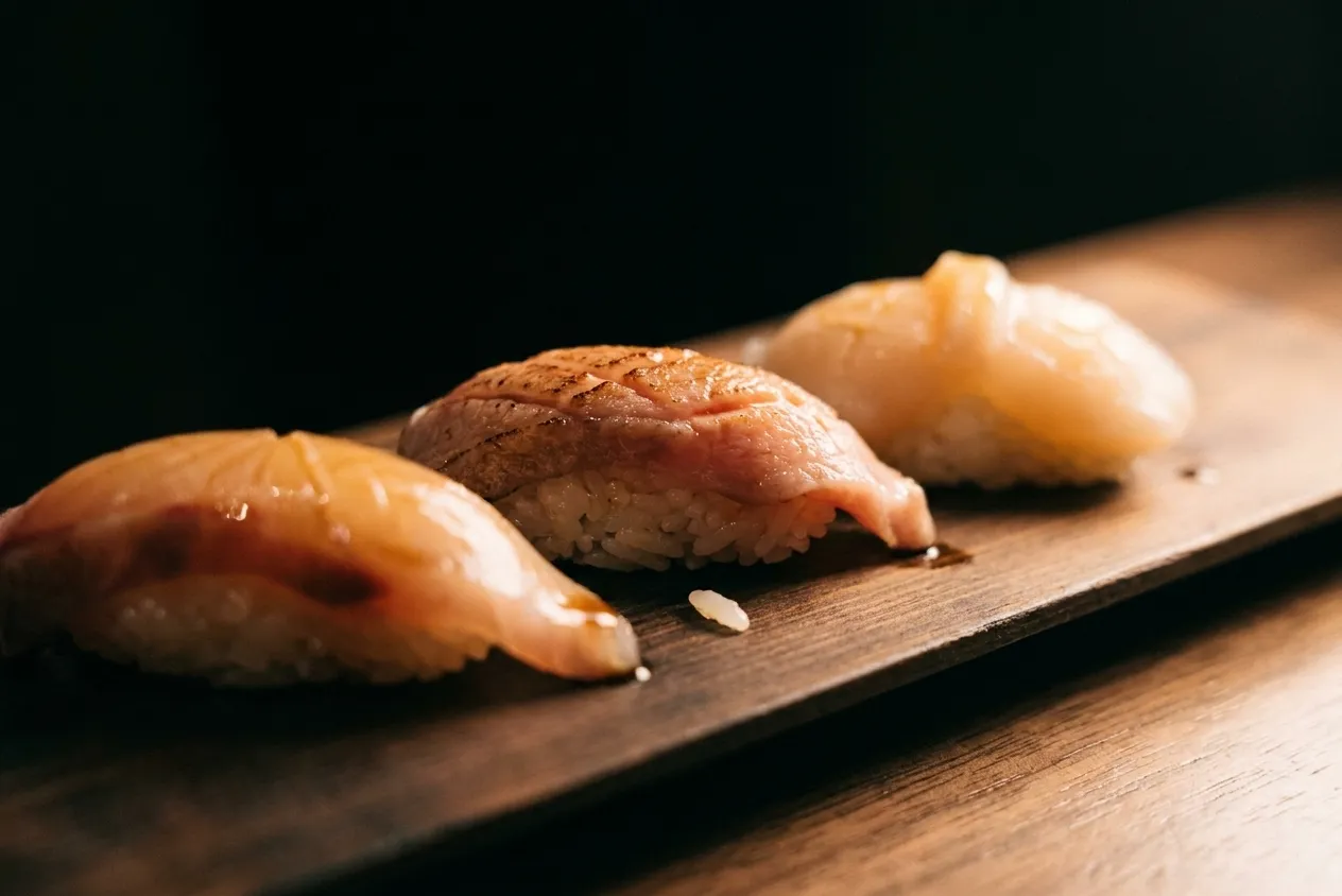 Three pieces of nigiri sushi lined up on a dark wooden slab under a single warm light, shot close and low with the background falling to black