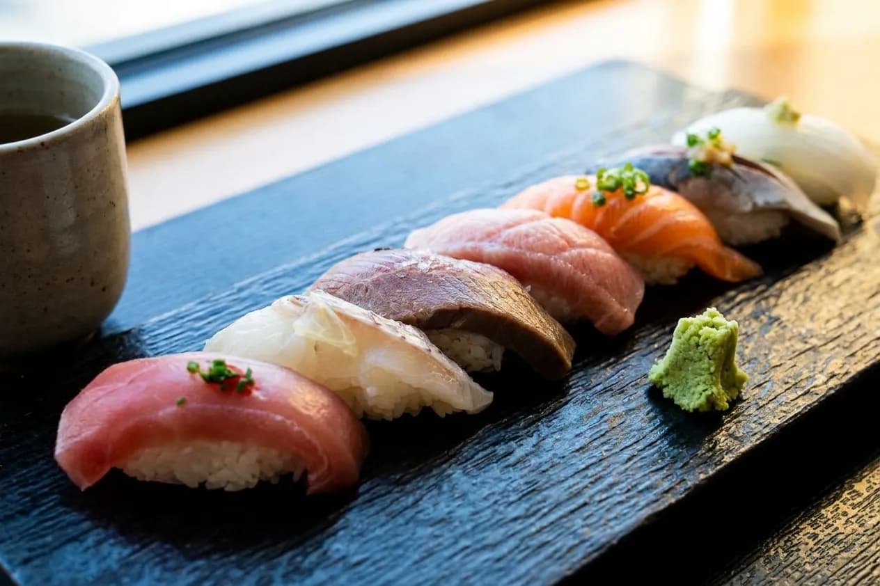 Eight pieces of seasonal nigiri lined up on a dark lacquered slab beside a plain ceramic tea cup, lit by soft window daylight