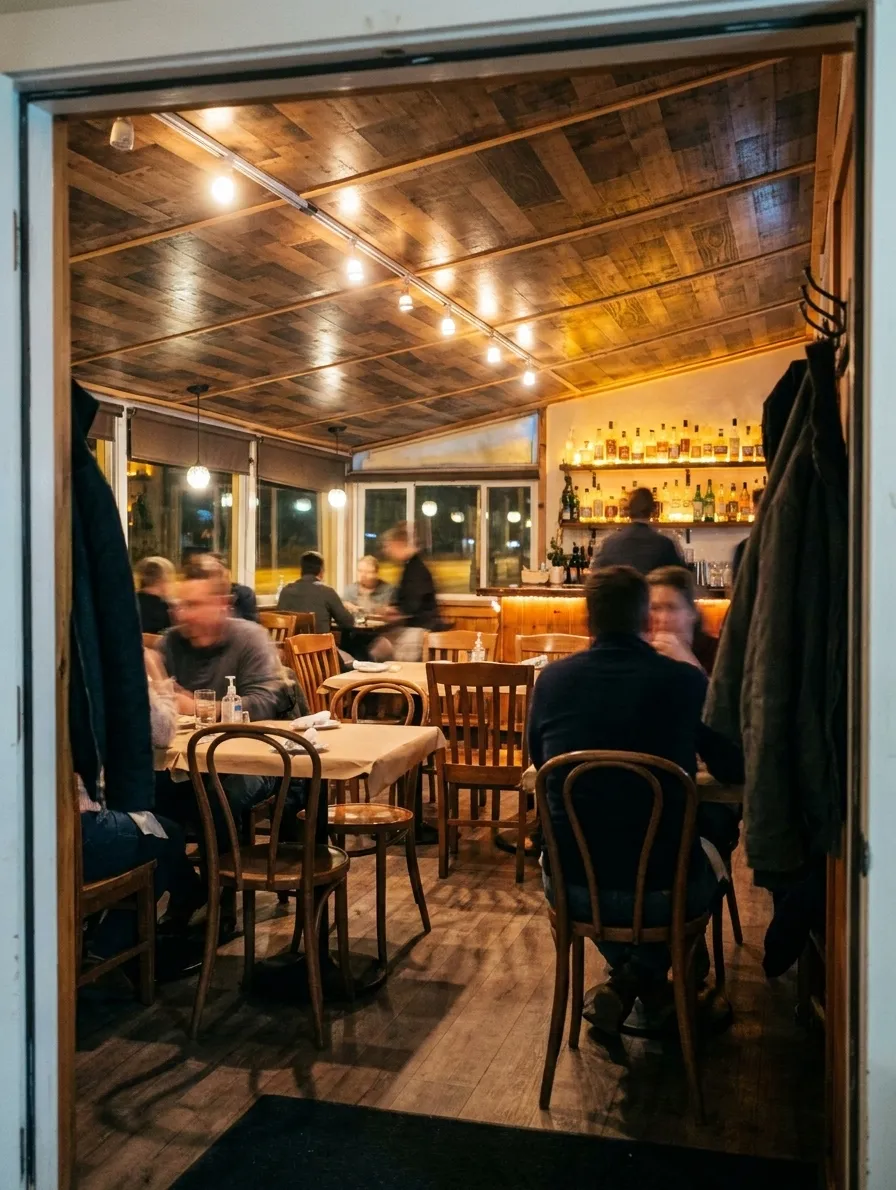 A warm, full restaurant dining room seen from just inside the doorway on a weeknight, with bentwood chairs, paper-topped tables and a lit bar along the back wall