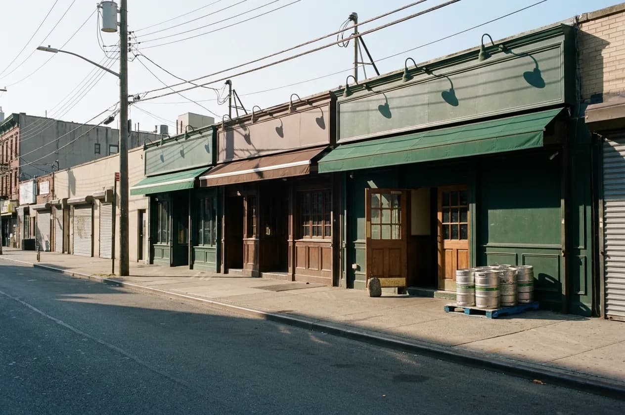 A wide Manhattan side street on a Saturday morning, a row of low pub frontages along the right-hand side, awnings still rolled up, early sun raking down the block