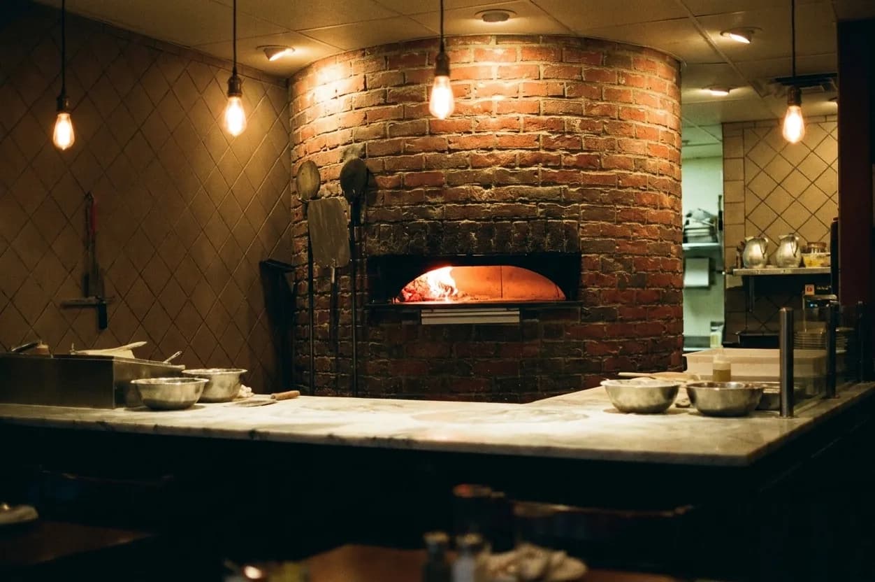 The open kitchen end of a neighbourhood pizzeria seen from the dining room, with a domed brick wood-fired oven glowing orange, steel peels leaning against the wall and warm pendant bulbs overhead
