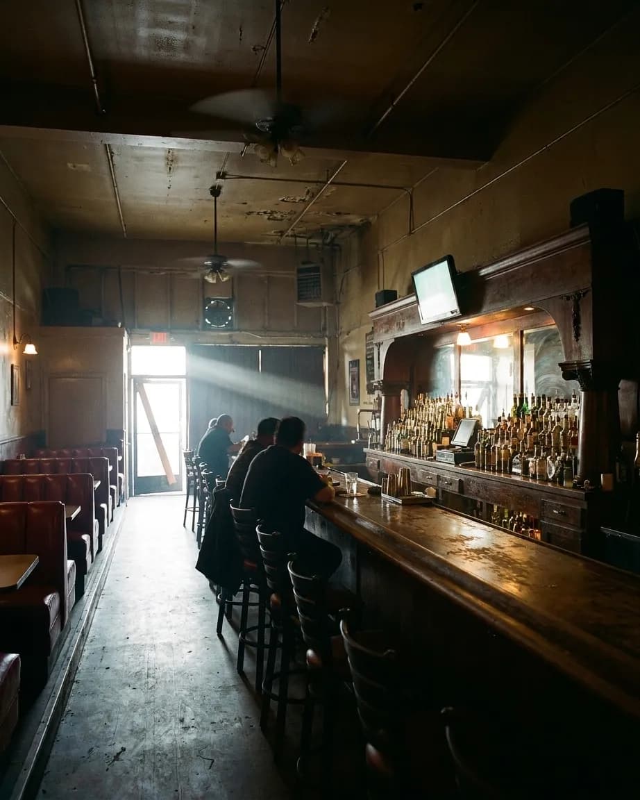 A dim eastside Los Angeles bar in the afternoon, red vinyl booths along one wall, a single screen glowing above the bar