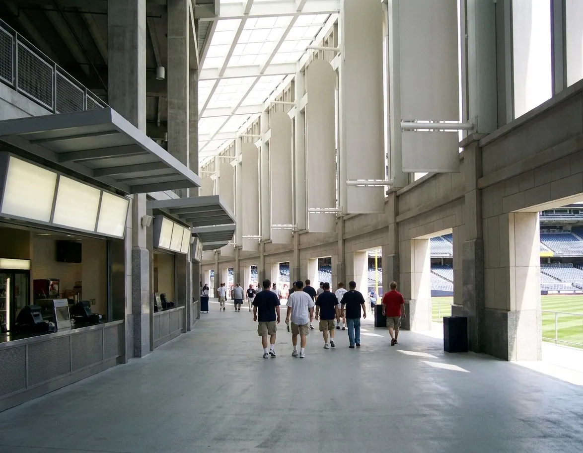 A ballpark concourse before a game, a curving walkway with concession openings on the left and a bright gap onto the seating bowl on the right