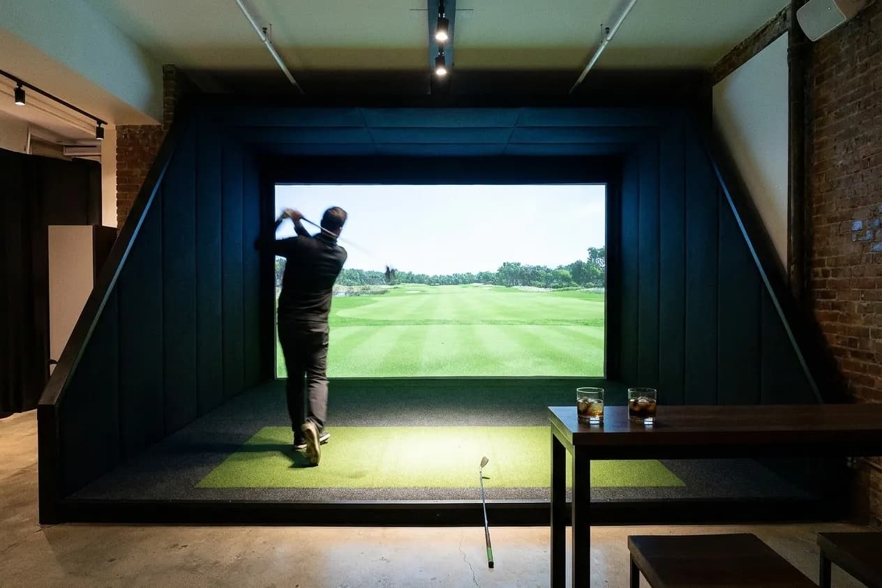 An indoor golf bay from behind the hitting area, a bright projection screen filling the far wall, dark padded side walls closing in toward it