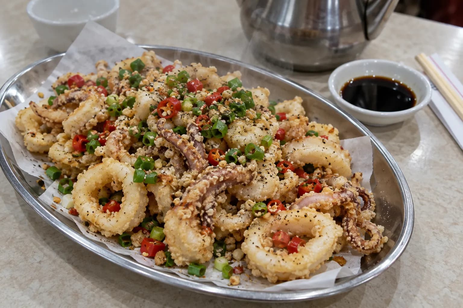 A metal dish of crisp fried squid with chilli and spring onion under bright light