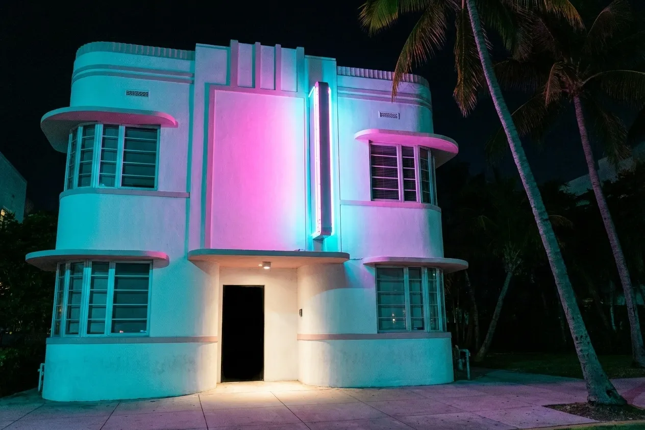 An Art Deco building facade on a Miami Beach street at night, curved corners and horizontal banding picked out in coloured light