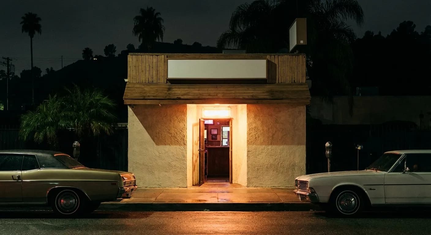 A tiny freestanding bar on a Los Angeles boulevard at night, a single doorway under a low overhang, neon glow spilling onto parked cars