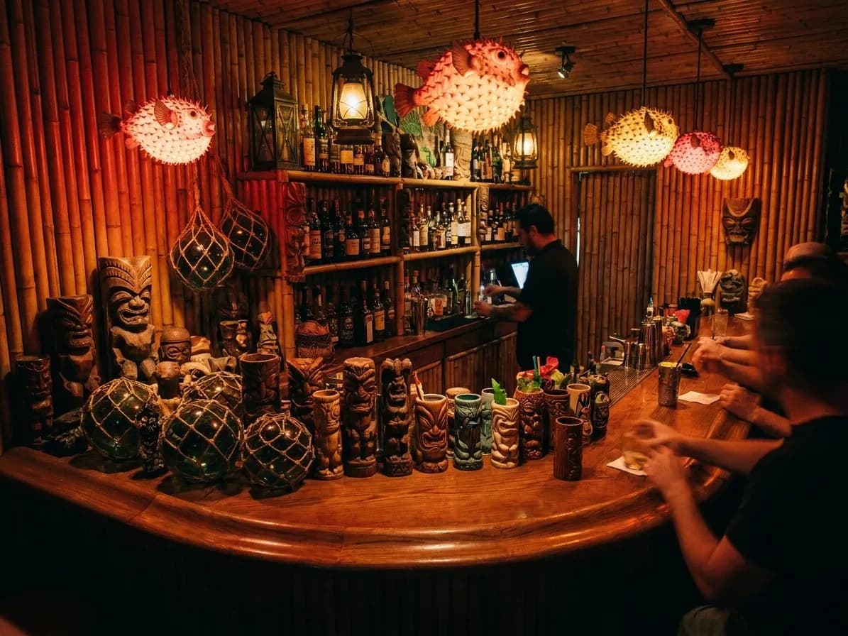 A cramped twelve-seat bar interior packed with carved figures, glass floats and hanging lamps, every surface covered, one bartender working the well