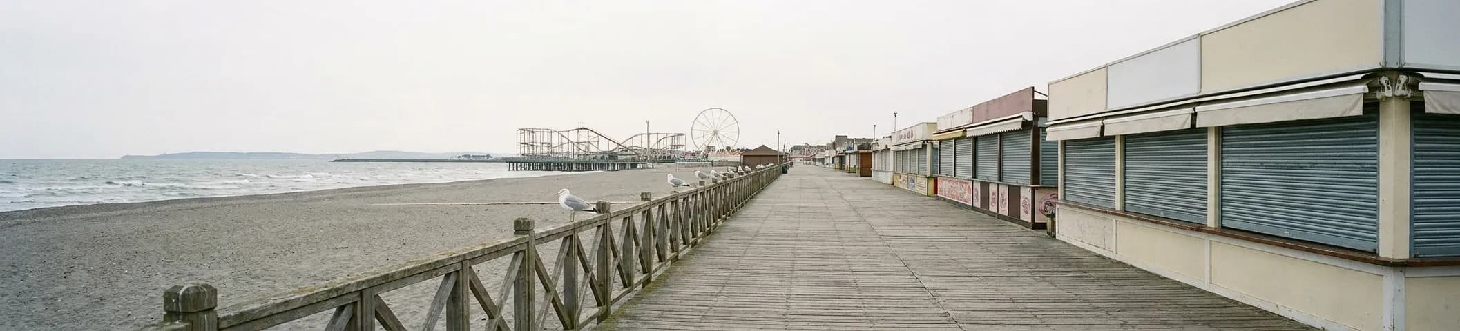 A wide wooden boardwalk running into the distance beside a beach, low food buildings along the inland side, a pale overcast sky above