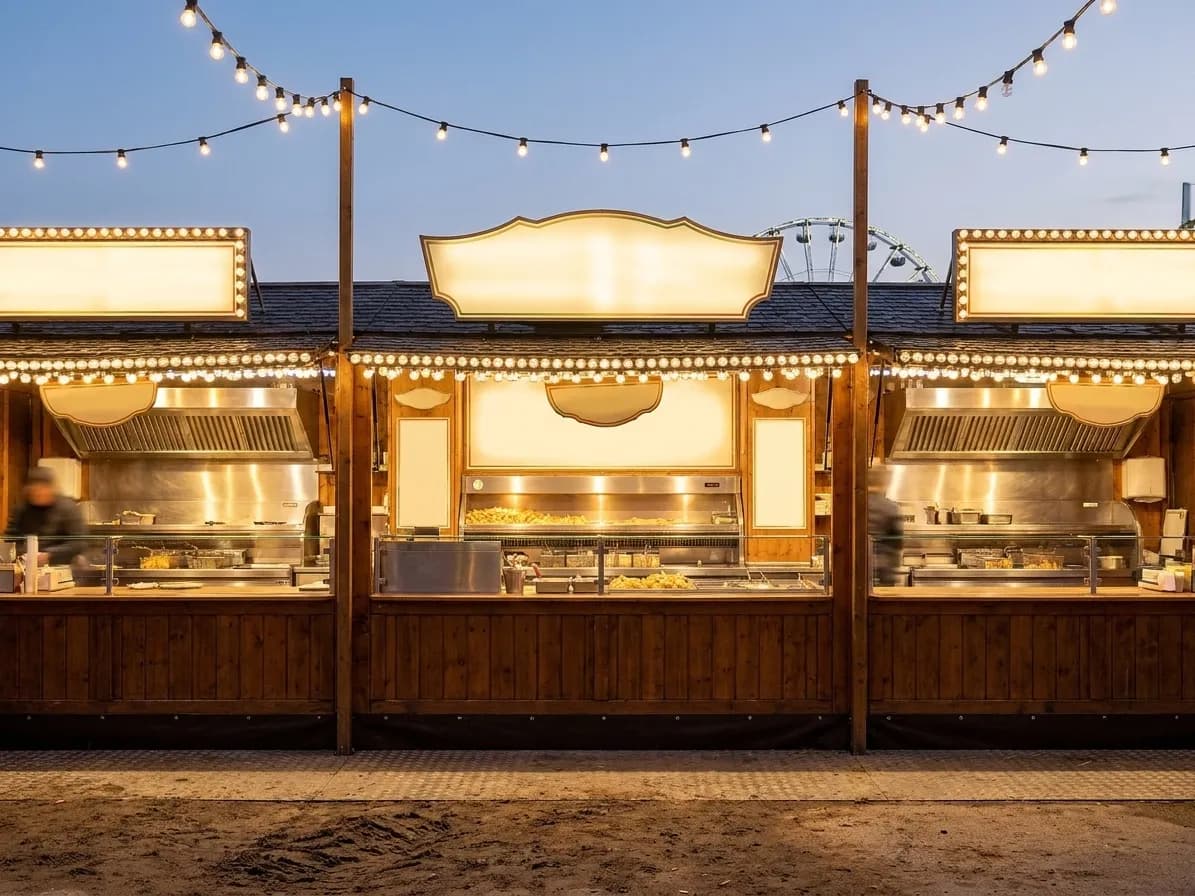 A row of carnival food stalls at dusk, blank illuminated sign panels above the counters, string lights overhead and a churned dirt walkway in front