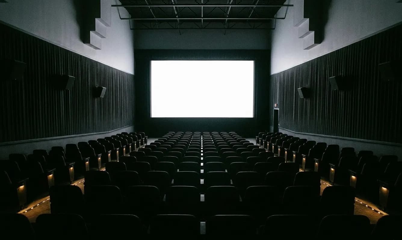 A small cinema auditorium seen from the back row, a blank illuminated screen at the far end, raked seating in shadow between