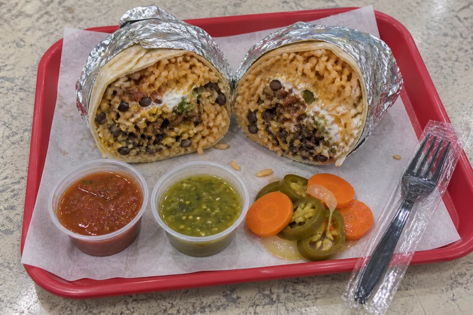 A large foil-wrapped burrito cut in half on a paper-lined tray under bright fluorescent light