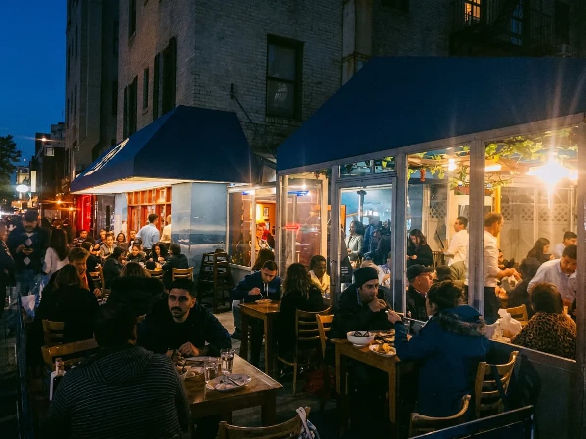 Night exterior of a Greek seafood taverna on a Ditmars Boulevard corner in Astoria, with blue awnings and crowded sidewalk tables