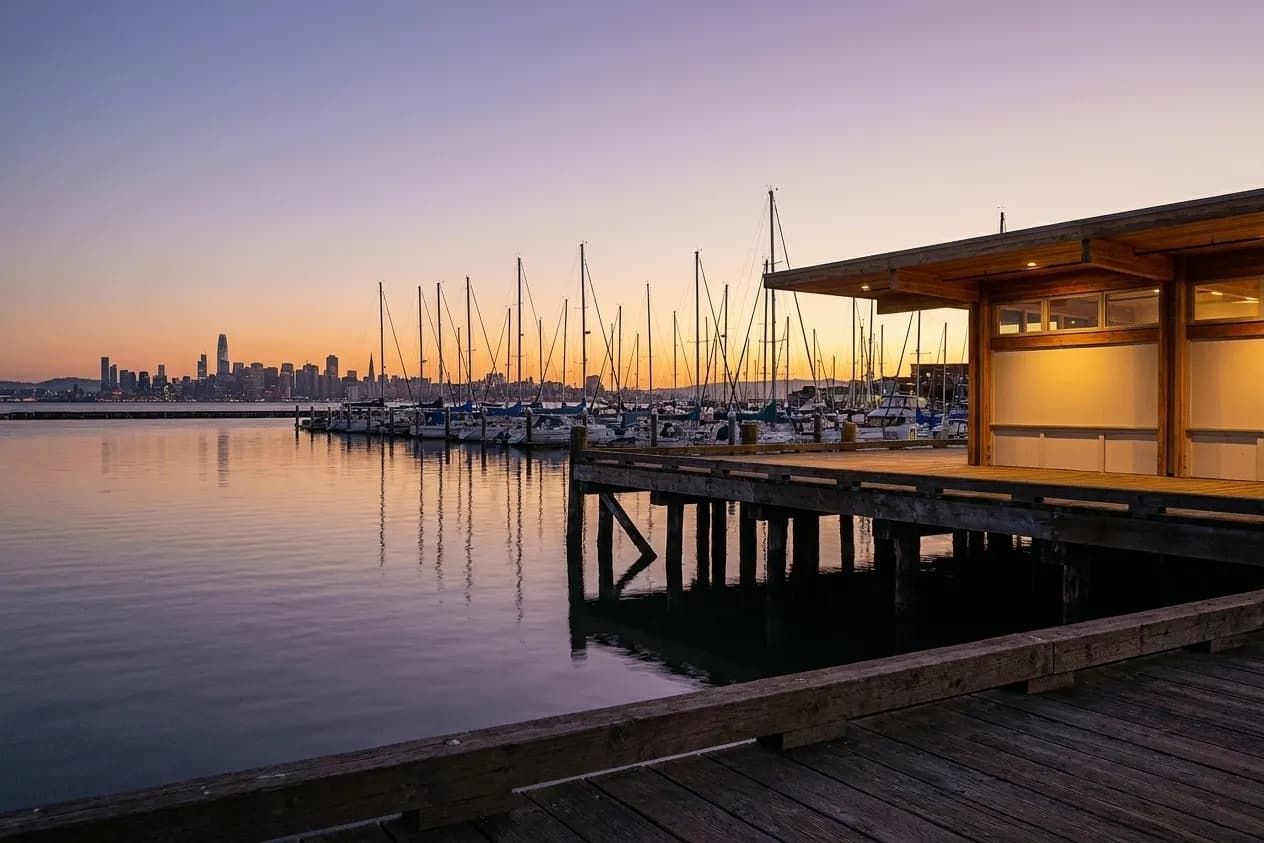 An East Bay waterfront at dusk, a low building along the shoreline, masts in a marina and the city skyline across the water