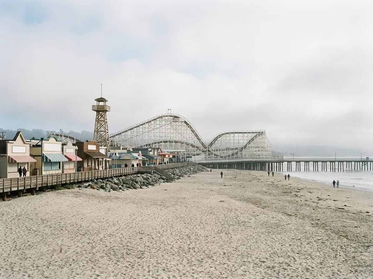 A seaside amusement boardwalk seen along the sand, a wooden coaster structure behind and a long pier reaching into the bay