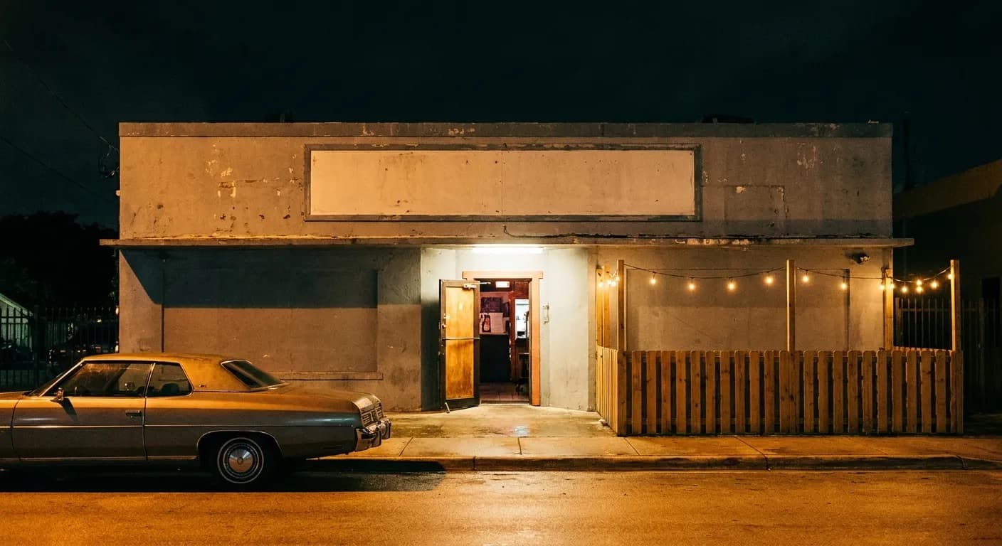 A painted dive-bar frontage at night in Miami, a lit doorway and a patio fence to one side