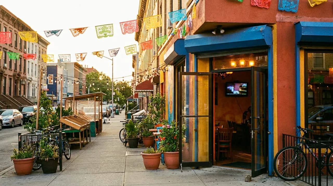 Vibrant NYC Mexican cantina exterior on a sunny weekend afternoon in Sunset Park Brooklyn, warm golden-hour sunlight, colorful papel picado banners across the sidewalk, open glass door with warm glow inside, small TV visible deep in the room.