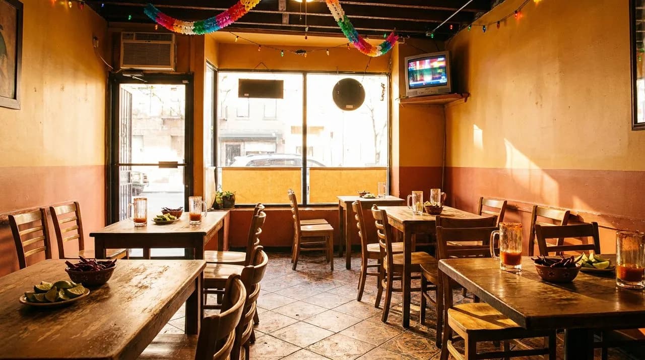 Bright cheerful interior of a NYC Liga MX cantina mid-afternoon, warm sunlight through the front window, tile floor, wooden tables with empty michelada glasses and lime wedges, small TV mounted in the corner glowing, festive busy atmosphere.