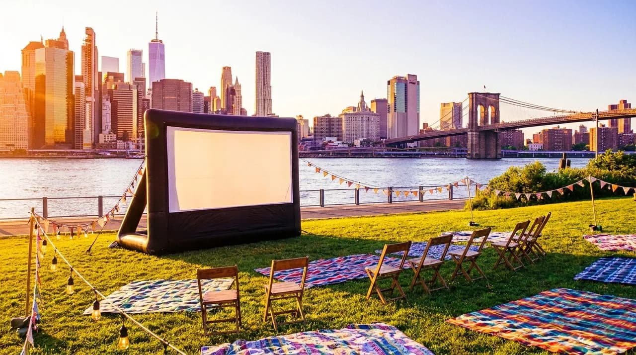 Brooklyn Bridge Park's Pier 1 lawn at late afternoon with a blank inflatable projector screen on grass, Manhattan skyline across the East River, and picnic blankets.