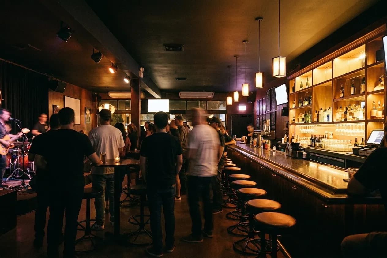 A busy bar room during a live set, people standing between tables, warm light from behind the bar