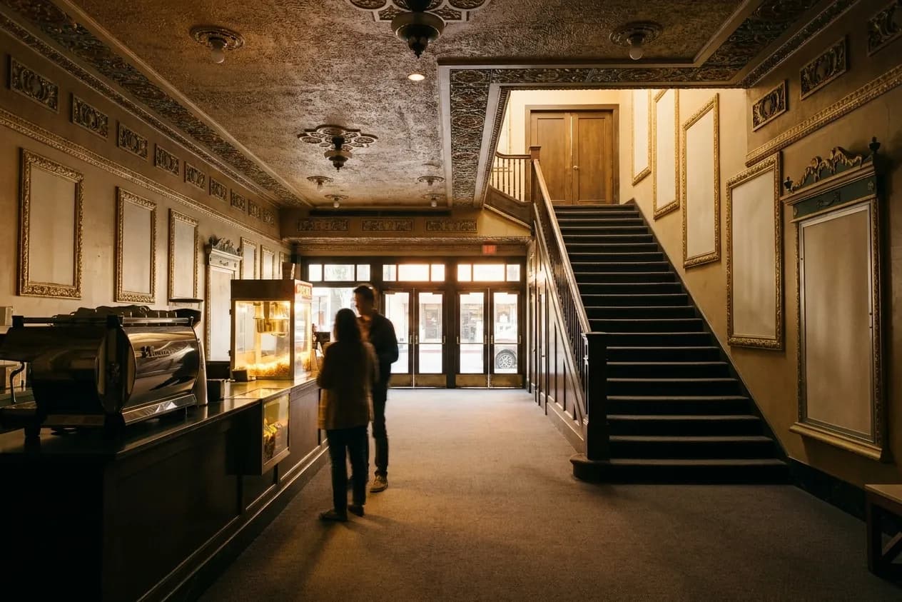 A cinema lobby in the evening, a concession counter to one side and a staircase rising toward the auditorium doors