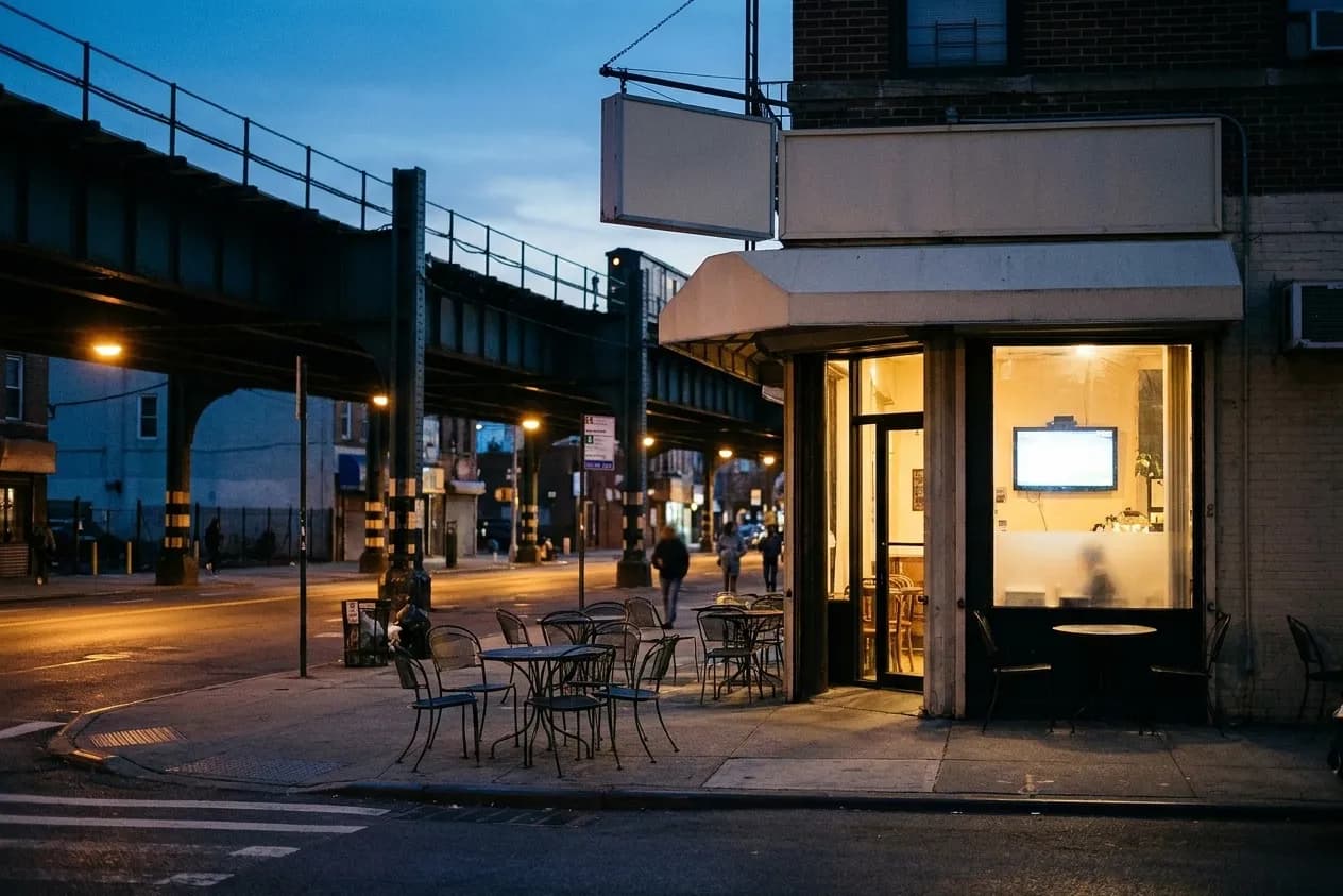 A Queens street corner at dusk with cafe tables outside and a screen visible through a window