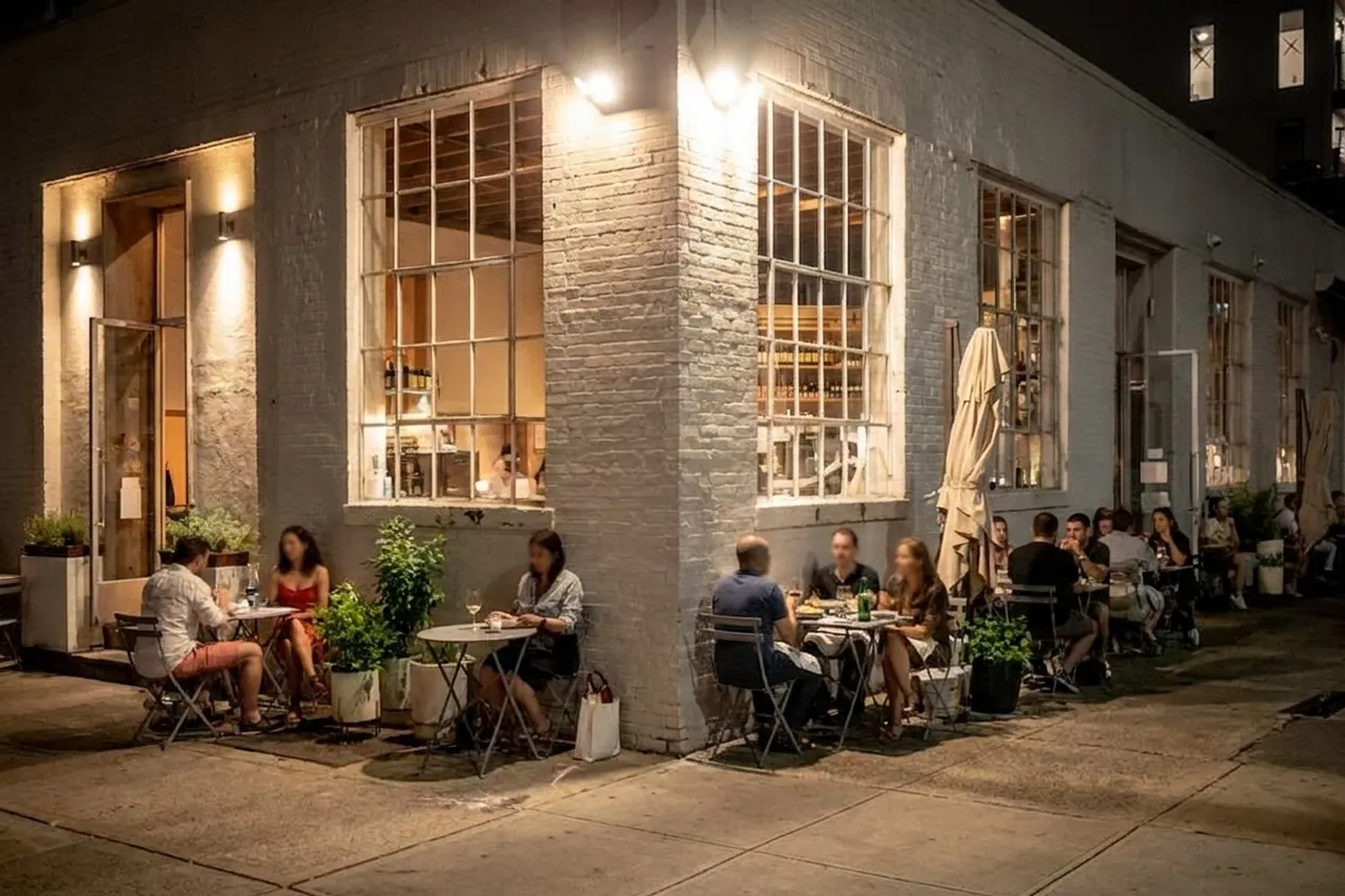 Night exterior of a white-brick corner restaurant with steel-framed windows glowing warm and couples at sidewalk tables