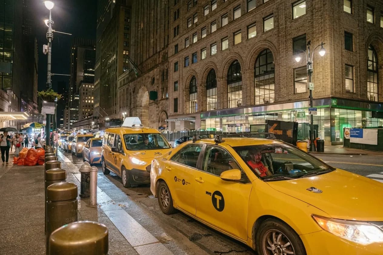 Yellow taxis queued on a New York City street at night with lit storefronts behind