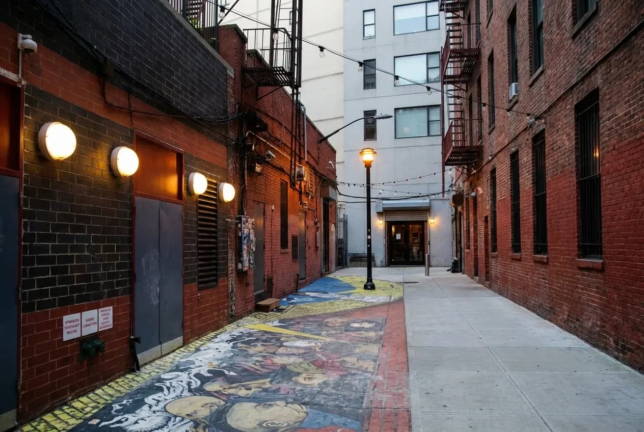 Brick-lined Extra Place alley in the East Village at dusk, string lights glowing above a painted pavement mural leading to a restaurant doorway