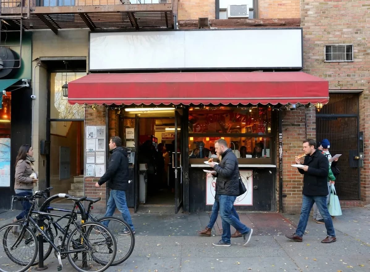 Pedestrians carrying pizza slices past a red-awning slice shop on a Greenwich Village street