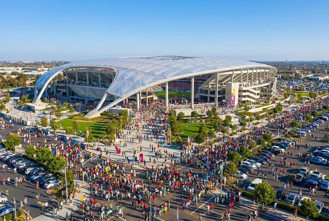 The sweeping modern curves of SoFi Stadium's exterior gleaming under a brilliant blue California sky, with palm trees and fans in World Cup colors streaming toward the entrance.