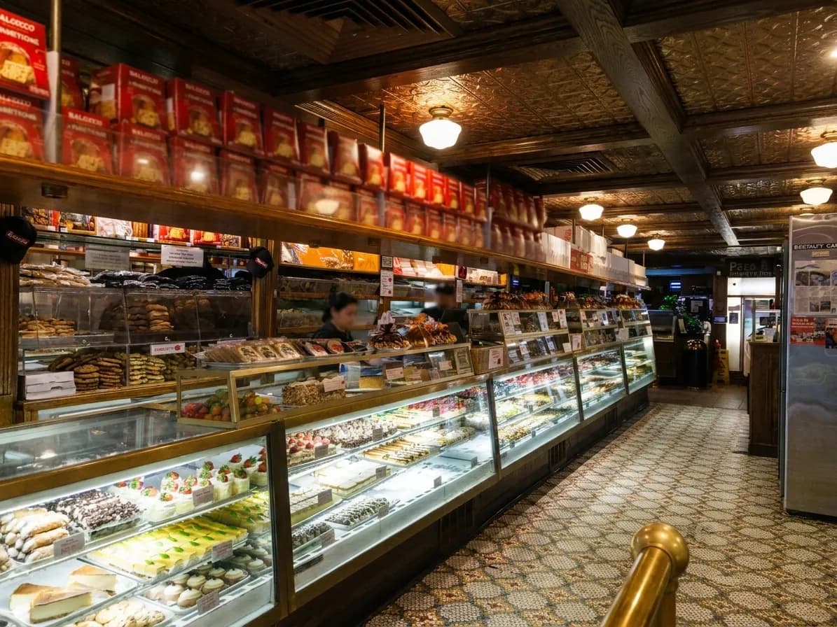 Warmly lit pastry counter with long glass display cases of cakes and cannoli under a pressed-tin ceiling in an old New York pasticceria