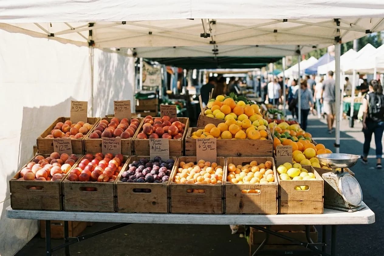 A Southern California farmers' market stall with crates of stone fruit and citrus under a canvas canopy in hard sun