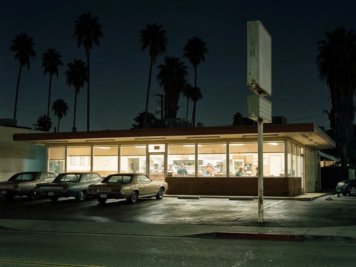 A low-slung Los Angeles diner at 3am with a long band of lit windows, an empty parking apron and a blank sign pole