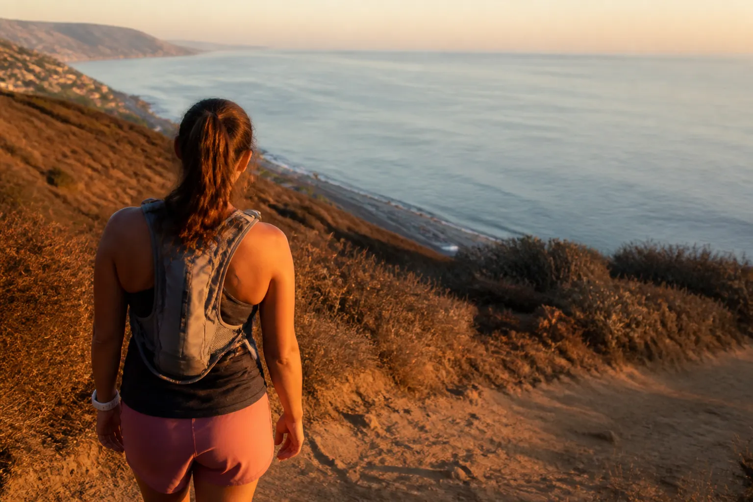 A hiker wearing a backpack looking out at the ocean from a scenic trail in Los Angeles during sunset.