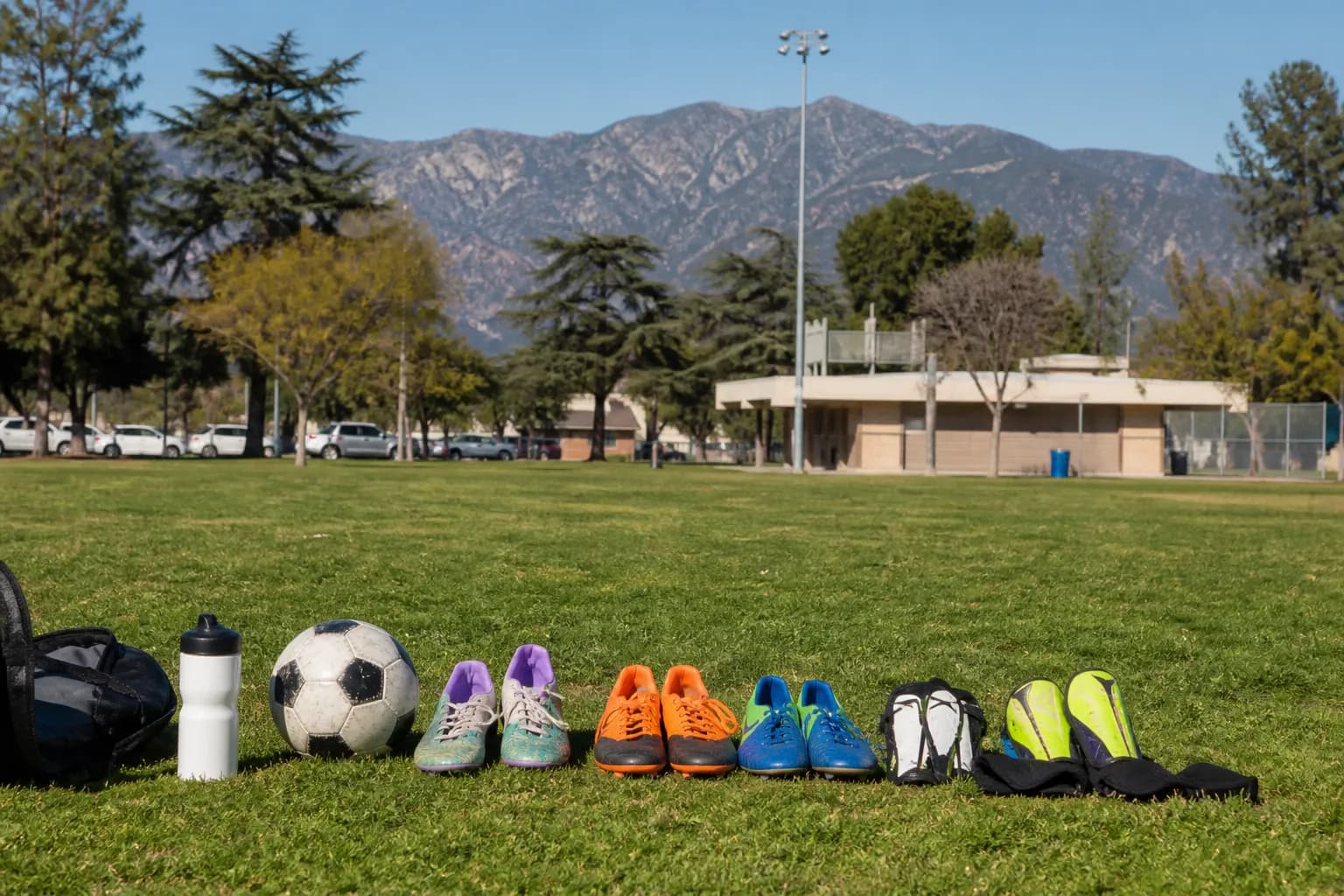 A collection of kids soccer gear including cleats, a soccer ball, and a water bottle resting on green grass.