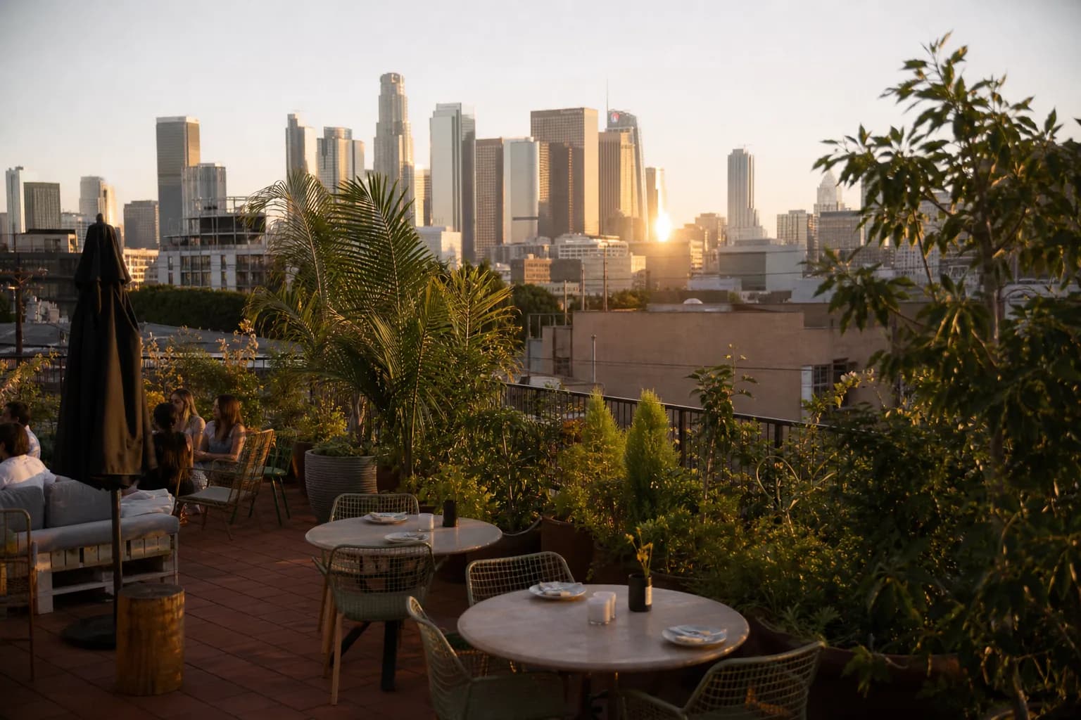 A wide landscape shot of a stylish rooftop lounge in Downtown Los Angeles during sunset, featuring warm orange skies reflecting off modern glass skyscrapers.