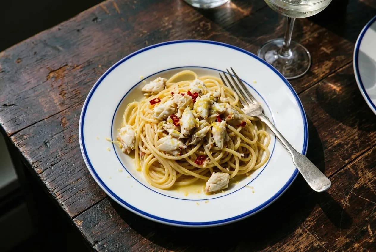 Overhead view of crab spaghetti with chili and lemon on white plate with fork on dark wood table in natural light.
