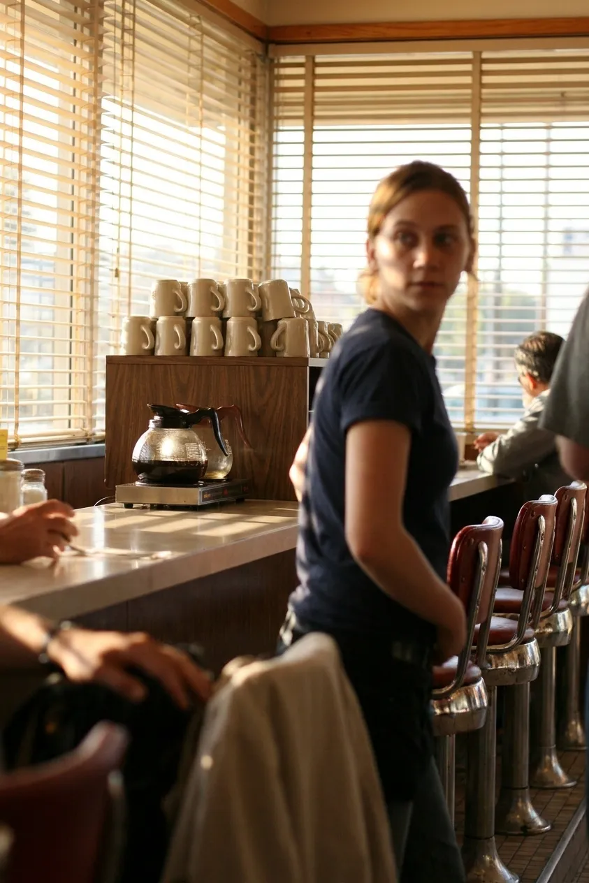 A Los Angeles diner counter in the morning with a row of chrome stools, a glass coffee pot on a warming plate and mugs upturned on a shelf