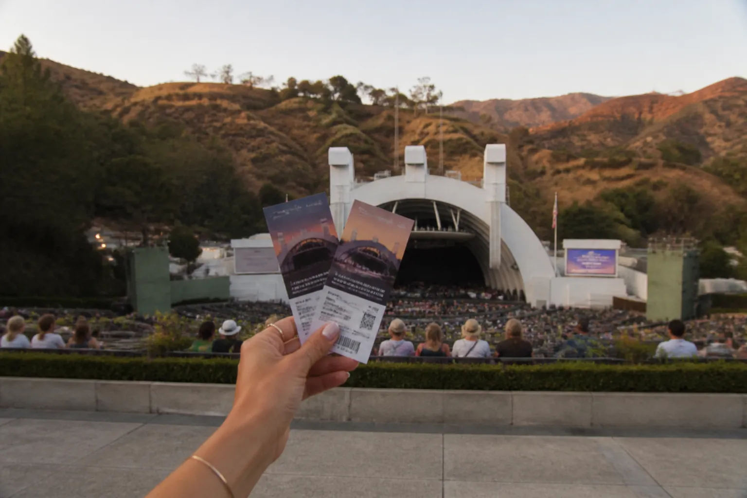 A close-up of physical concert tickets held up against the backdrop of the Hollywood Bowl amphitheater structures.