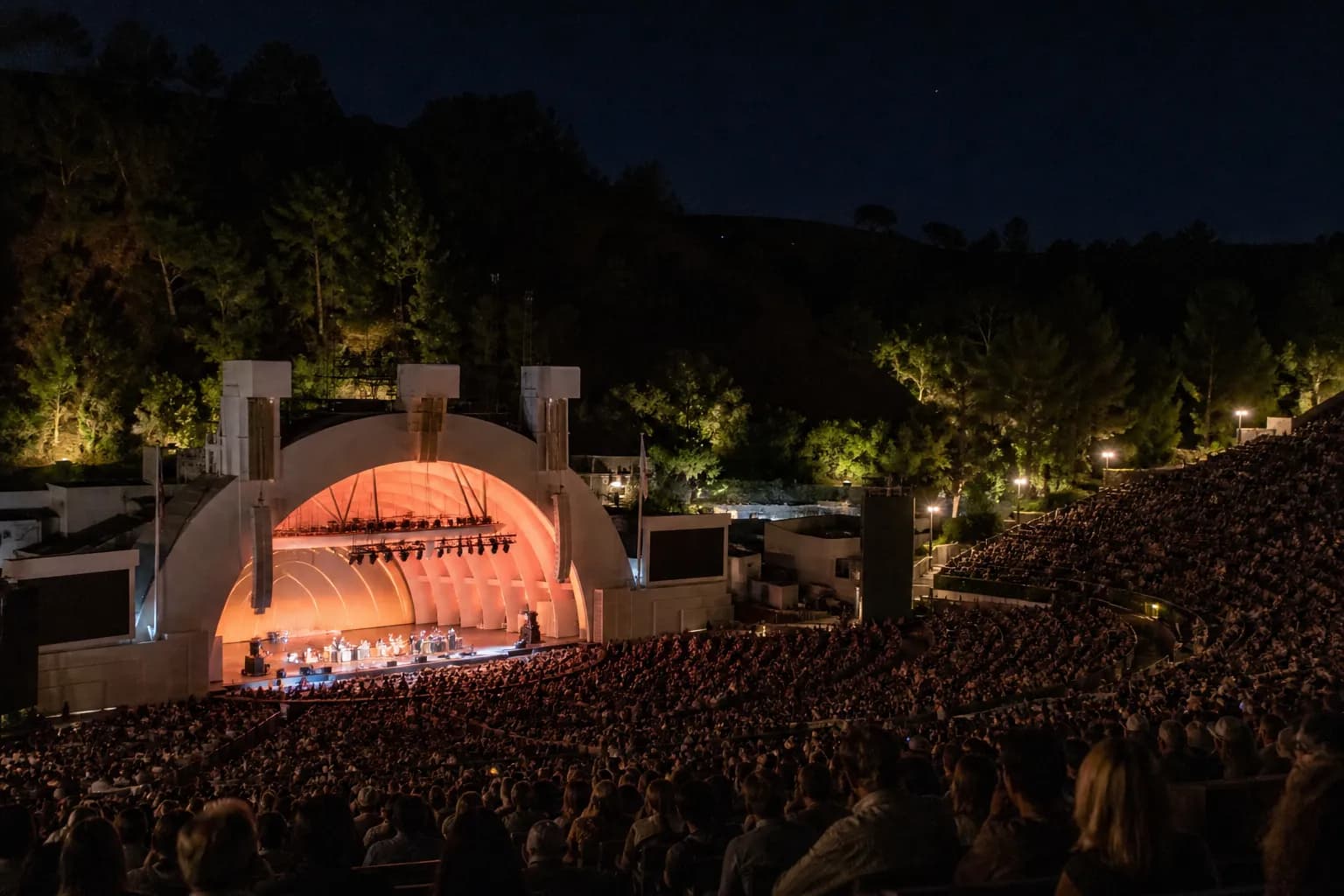 The illuminated stage of the Greek Theatre surrounded by dark pine trees in Griffith Park at night.