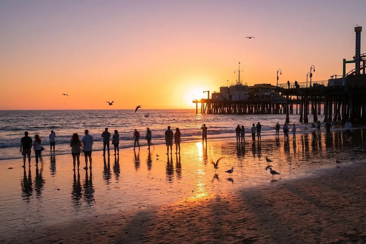 A Los Angeles beach at sunset with people scattered along the sand facing the water and a pier silhouette to one side