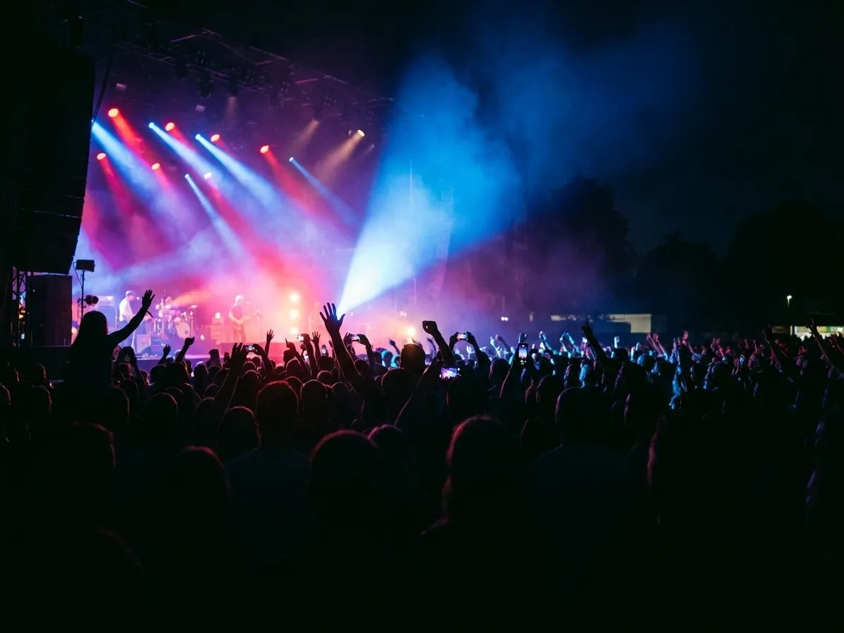 A gig crowd from behind with raised arms and phone screens silhouetted against a bright stage and coloured beams cutting through haze